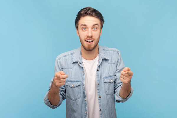 Портрет удивленного шокированного удивленного небритого мужчины указывает пальцем в камеру, видит что-то невероятное спереди, в джинсовой рубашке. Внутренний снимок изолирован на синем фоне.