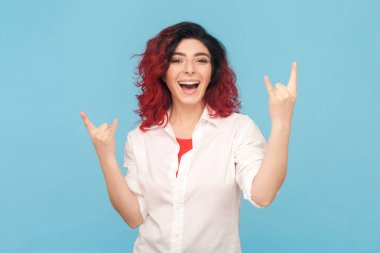 Havalı kızıl saçlı, çekici bir kadının portresi rock 'n roll jesti yapıyor. Keyfi yerinde, beyaz gömlek giyiyor. Mavi arka planda kapalı stüdyo çekimi.