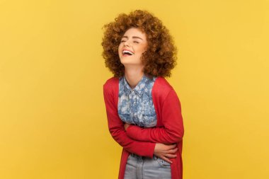 Afro saç stili, neşeli bir kadın ellerini karnının üstünde tutarak kahkahalarla gülüyor. Komik anekdotlar duyduktan sonra gülmeden duramıyor. Sarı arka planda kapalı stüdyo çekimleri.