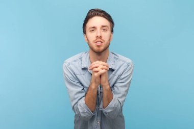 Umutsuz, mutsuz, tıraşsız, af dileyen, dudaklarını çantasına dayayan, avuç içini dua eden, merhamet dileyen ve yardımınıza ihtiyacı olduğunu söyleyen bir adam. Mavi arkaplanda kapalı çekim.