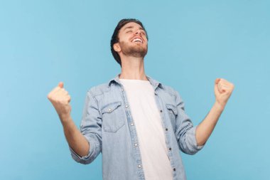 Adam yumruklarını sıkar, heyecanla bağırır, tatlı bir başarıyla sevinir, zaferin tadını hisseder, en sevdiği takım için bağırır, kot gömlek giyer. Mavi arkaplanda kapalı çekim.