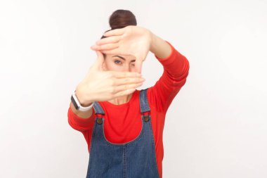 Portrait of concentrated woman photographer with hair bun looking through frame of fingers, creates new picture, wearing denim overalls. Indoor studio shot isolated on white background