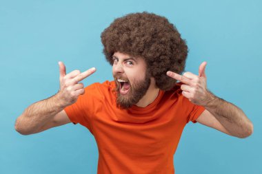 Orta parmaklarını gösteren turuncu tişörtlü, Afro saçlı ve negatif ve saygısız tavırları olan öfkeli bir adamın portresi. Mavi arka planda kapalı stüdyo çekimi.