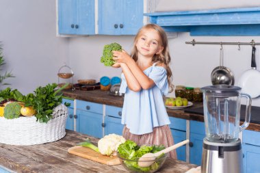 Mavi elbiseli, elinde yeşil brokoliyle kameraya bakıp gülümseyerek modern mutfakta sebze salatası hazırlayan sevimli küçük bir kızın portresi..
