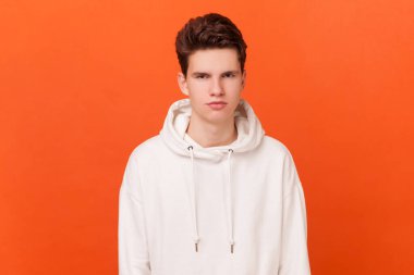 Beyaz kapüşonlu, ciddi ve yakışıklı bir adamın portresi. Kameraya bakıyor, sinirli ve agresif. Turuncu arka planda kapalı stüdyo çekimleri.