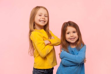 İki neşeli, mutlu küçük kızın portresi. Elleri bağlı, mutlu bir şekilde duruyorlar. Mutluluk ve neşeyi ifade ediyorlar. Pembe arka planda kapalı stüdyo çekimleri.