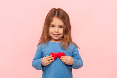 Sevimli kahverengi saçlı küçük kızın portresi kameraya bakıyor, küçük kırmızı kalpler gösteriyor, aşkı ifade ediyor, mavi kazak giyiyor. Pembe arka planda kapalı stüdyo çekimleri.