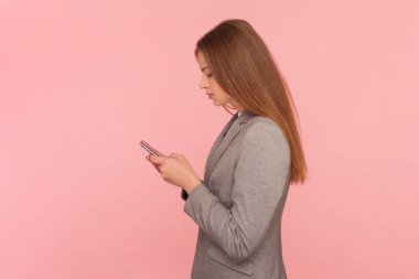 Kahverengi saçlı, cep telefonu kullanan, mesaj yazan, internette sörf yapan, takım elbise giyen ciddi bir kadının yan görünüm portresi. Pembe arka planda kapalı stüdyo çekimleri.