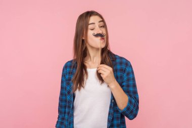 Eğlenceli, kahverengi saçlı, kağıt bıyıklı, neşeli bir şekilde kameraya göz kırpan ve kareli gömlek giyen bir kadının portresi. Pembe arka planda kapalı stüdyo çekimleri.