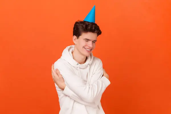 stock image Happy birthday to me. Cute man in white hoodie and blue party cone embracing himself with expression of pleasure, happy to celebrate holiday alone. Indoor studio shot isolated on orange background.