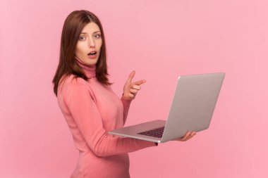 Kahverengi saçlı, laptopta çalışan, şaşkın suratlı, balıkçı yaka giyen bir kadının yan görünüm portresi. Pembe arka planda kapalı stüdyo çekimleri