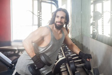Kendine güvenen, uzun kıvırcık saçlı, spor salonunda tek bir dambılı tutan, pazı ve kol kasları için egzersiz yapan kendine güvenen yakışıklı bir adamın portresi. Kapalı çekim.