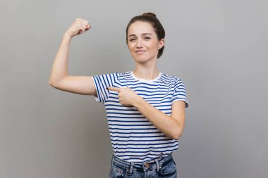 I am strong. Portrait of woman wearing striped T-shirt raising hand showing biceps, looking at camera with confidence pride, feeling power. Indoor studio shot isolated on gray background. clipart