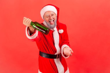 Happy funny elderly man with gray beard wearing santa claus costume standing with alcohol bottle and offering beer, celebrating new year. Indoor studio shot isolated on red background. clipart