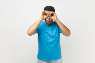 Portrait of unshaven man standing making glasses shape with fingers, looking through binoculars hand gesture and expressing shock, astonishment. Indoor studio shot isolated on gray background. clipart