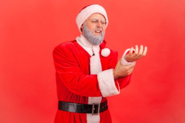 Elderly man with gray beard wearing santa claus costume standing with grimace of pain, massaging sore wrist, suffering hand injury or sprain. Indoor studio shot isolated on red background. clipart