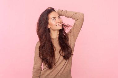 Attractive dreaming smiling woman with wavy hair, keeps hand on her head, looking away, expressing positive emotions, wearing wearing brown pullover. Indoor studio shot isolated on pink background clipart