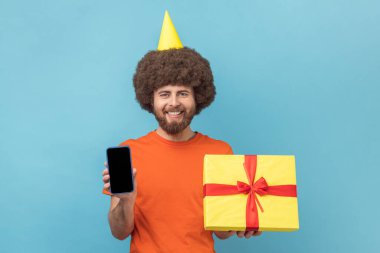 Afro saçlı, turuncu tişörtlü ve parti konisi taşıyan hediye kutusu ve cep telefonlu bir adam. Reklam için fotokopi alanı, boş ekran. Mavi arka planda kapalı stüdyo çekimi.