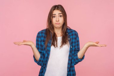 Kafası karışmış kahverengi saçlı kadının portresi omuz silkip kameraya belirsiz bir bakışla bakıyor, kareli gömlek giyiyor. Pembe arka planda kapalı stüdyo çekimleri.