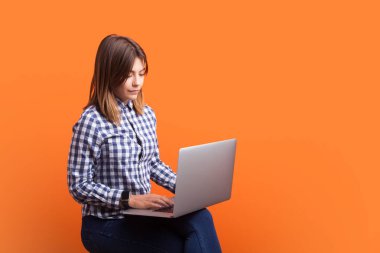 Ciddi bir kadın portresi, kahverengi saçlı, dizüstü bilgisayarıyla oturan ve uzaktan çalışan, uzaktan kumandalı, kareli gömlek giyen. Turuncu arka planda kapalı stüdyo çekimleri