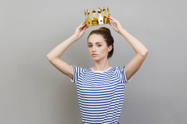 Портрет серьезной уверенной женщины в полосатой футболке и золотой короне, выглядящей с высокомерием и уверенностью, привилегированным статусом. Крытая студия, снятая на сером фоне.