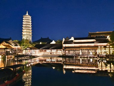 Güzel aydınlatılmış bir pagoda ve geleneksel Çin binaları derin mavi gökyüzünün altındaki sakin suları yansıtır. 
