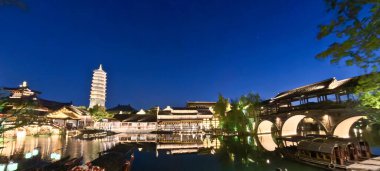 Güzel aydınlatılmış bir pagoda ve geleneksel Çin binaları derin mavi gökyüzünün altındaki sakin suları yansıtır. 