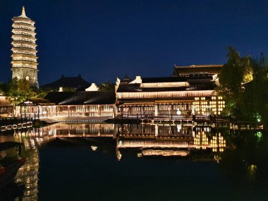 Güzel aydınlatılmış bir pagoda ve geleneksel Çin binaları derin mavi gökyüzünün altındaki sakin suları yansıtır. 
