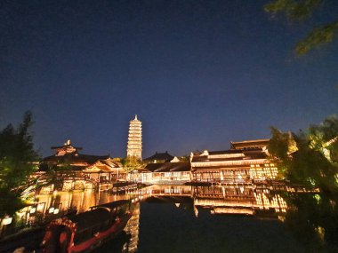 Güzel aydınlatılmış bir pagoda ve geleneksel Çin binaları derin mavi gökyüzünün altındaki sakin suları yansıtır. 