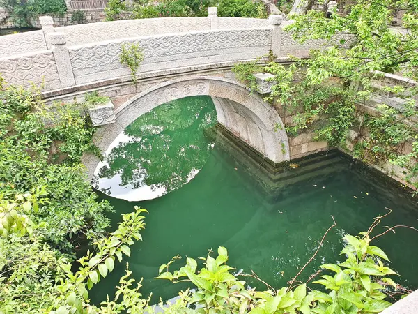 Geleneksel taş kemer köprüsü, tarihi Çin binaları ve yemyeşil ağaçlar ve bitkilerle çevrili sakin bir kanalı kapsar. 