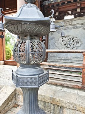 Karmaşık oymaları ve asılı çanları olan süslü bronz bir fener geleneksel Çin yardım sanatı ile süslenmiş taş bir duvarın yanında duruyor. 