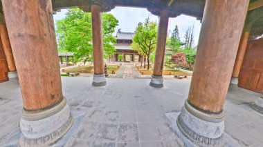 Geleneksel ahşap bir tapınak yapısının içinde, büyük oyulmuş sütunlar yüksek bir çatıyı destekliyor, ötesinde huzurlu bir bahçe manzarası oluşturuyor. 