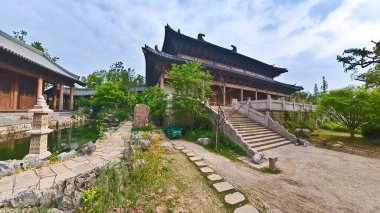 Geleneksel ahşap bir tapınak yapısının içinde, büyük oyulmuş sütunlar yüksek bir çatıyı destekliyor, ötesinde huzurlu bir bahçe manzarası oluşturuyor. 