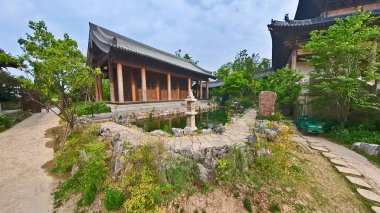 Geleneksel ahşap bir tapınak yapısının içinde, büyük oyulmuş sütunlar yüksek bir çatıyı destekliyor, ötesinde huzurlu bir bahçe manzarası oluşturuyor. 