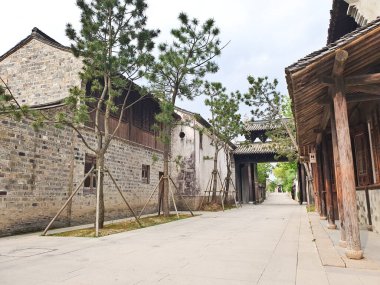 Geleneksel Çin köyü sahnesinde eski ahşap binalar, taştan kaldırımlar, kanallar ve bulutlu bir gökyüzünün altında yapraklı ağaçlar yer alıyor. 