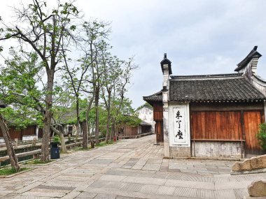 Geleneksel Çin köyü sahnesinde eski ahşap binalar, taştan kaldırımlar, kanallar ve bulutlu bir gökyüzünün altında yapraklı ağaçlar yer alıyor. 