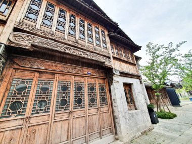 Geleneksel ahşap Çin binası. Karmaşık kafes pencereleri ve oymaları var. Gün ışığında ağaçlarla kaplı, sakin bir kaldırım taşı caddesi boyunca yer alıyor. 