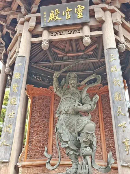 Vahşi bir savaşçı heykeli süslü ahşap bir köşkün altında duruyor. Geleneksel Çin mimari detayları ve altın Çin kaligrafisi yazıtları var. 