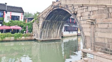 A traditional stone arch bridge spans a calm canal, with historic buildings, greenery, and tourists adding cultural charm. 