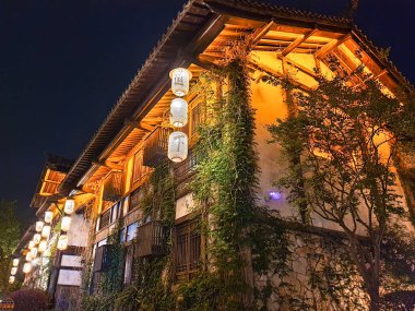 Geleneksel ahşap bina yoğun yeşil sarmaşıkla kaplı, dekoratif pencereler, ahşap kirişler ve berrak bir gökyüzü altında yemyeşil bitki örtüsü 