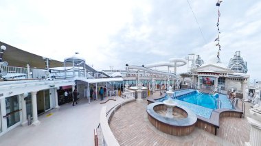 Geniş bir yüzme havuzu ve güneşli güvertesi olan sakin mavi bir denizde yelken açan büyük bir yolcu gemisinin panoramik görüntüsü.