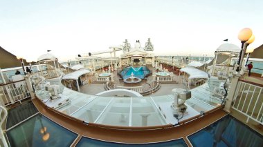 Geniş bir yüzme havuzu ve güneşli güvertesi olan sakin mavi bir denizde yelken açan büyük bir yolcu gemisinin panoramik görüntüsü.