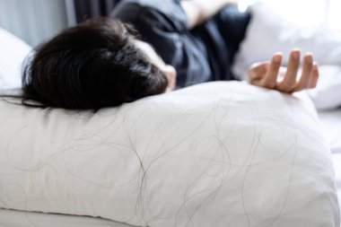 Yakın plan, yastıkta uzun siyah saç dökülmesi, saç dökülmesi veya kafa derisi sorunu, yatakta uyuyan Asyalı kadın, yatak odasında uzanan alopecia hastası, güzellik, sağlık bakımı ve tıbbi konsept.