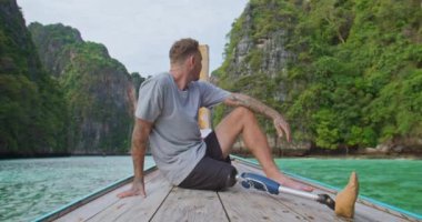 Uzun kuyruk teknesinin önünde oturan yapay biyonik bacaklı genç beyaz erkek Tayland 'ın Phi Adaları' nda yaz tatili sırasında harika bir deniz manzarası izliyor..