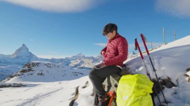 Genç Asyalı iş adamı karlı dağın tepesinde oturmuş dizüstü bilgisayarını kullanarak İsviçre 'de kışın ünlü Matterhorn arka planının üzerinde çalışıyor. Dijital Göçebe İnternet Teknolojisi.