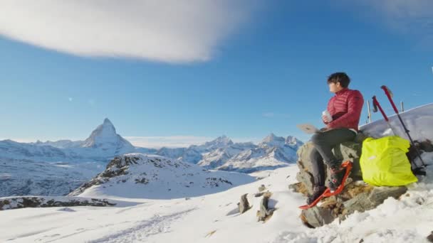 若いアジアのビジネスマンフリーランスは 冬の有名なマッターホルンの背景の上にスイスで旅行しながら リモートで作業ラップトップを使用して雪山の上に座っています デジタルノマドオンライン技術 — ストック動画