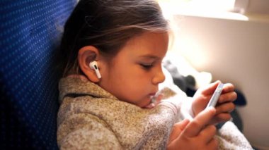 Trenle seyahat eden ve telefondaki kablosuz kulaklıklarla çizgi film izleyen kız çocuğu. Yüksek kalite fotoğraf
