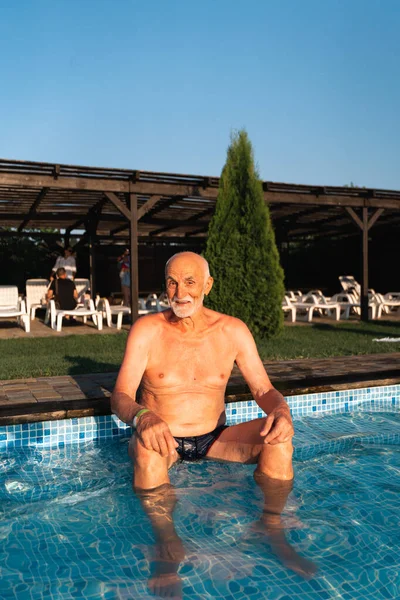 80 years old white man with beard relaxing near the pool on a hot summer sunny day. Lifestyle candid style, happy real emotions. High quality photo