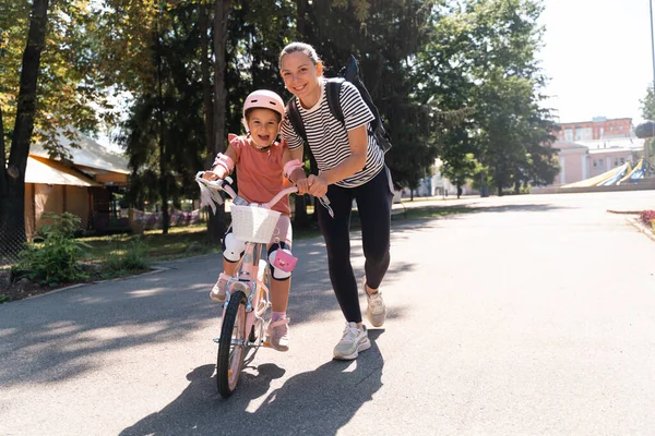 Mother learning child girl to ride a bike in the park in the summer. Happy quality family time together. Empowering, essential physical skills for kids. High quality photo
