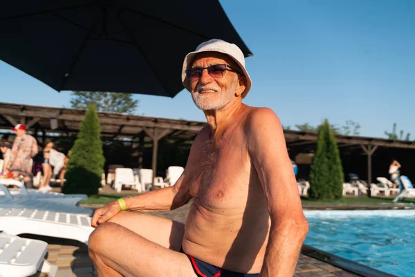 80 years old white man with beard relaxing near the pool on a hot summer sunny day. Lifestyle candid style, happy real emotions. High quality photo